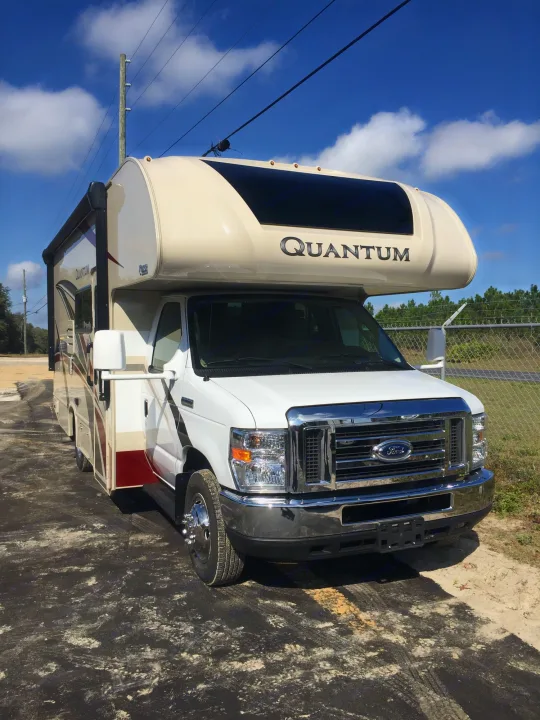 White and beige Ford Quantum RV rental parked on a partly paved lot under a clear sky with clouds.. Thor Motor Coach Quantum GR 22 2020