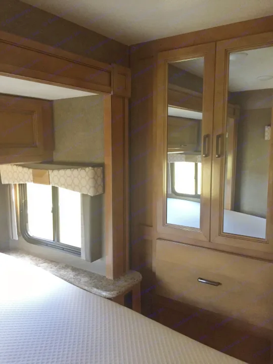 An RV rental bedroom showcasing a bed, window with a patterned valance, and mirrored wardrobe in a warm wood interior.. Thor Motor Coach Quantum GR 22 2020