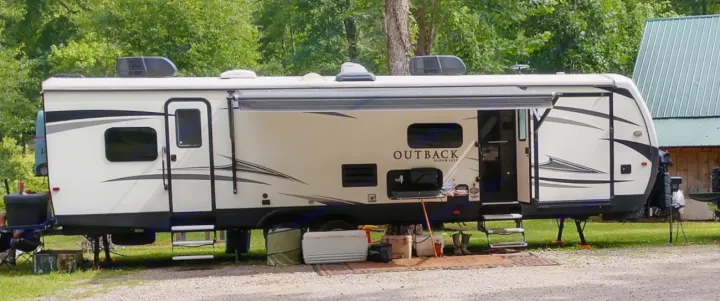 A white Outback travel trailer, likely an RV rental, is set up at a leafy campsite with its awning extended, steps down, and outdoor gear ready.. Keystone Outback 2018