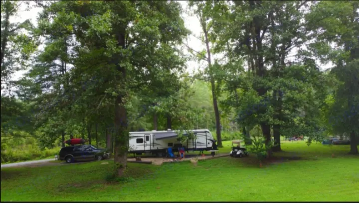 RV rental at a shaded campground among tall trees, with an SUV, kayak, and golf cart for relaxing outdoor fun.. Keystone Outback 2018