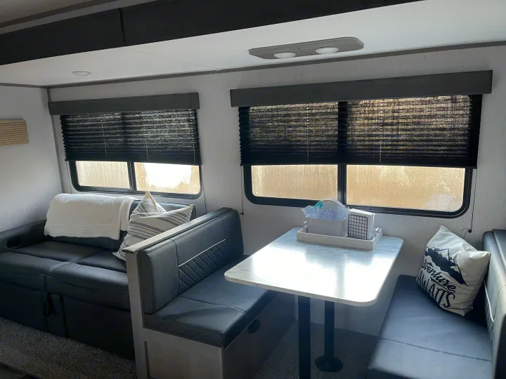 View inside an RV rental. Features dark gray leather-look sofa, dinette with white marble table, and black pleated window blinds.. Dutchmen Coleman 2022