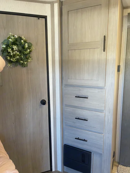 Modern RV rental interior: Light wood door with wreath, storage cabinets, and drawers with black hardware.. Dutchmen Coleman 2022
