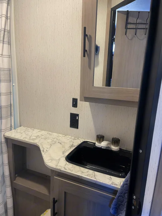 rv rental bathroom vanity area with a black sink, marble-patterned counter, mirror, and cabinet storage. Striped shower curtain.. Dutchmen Coleman 2022