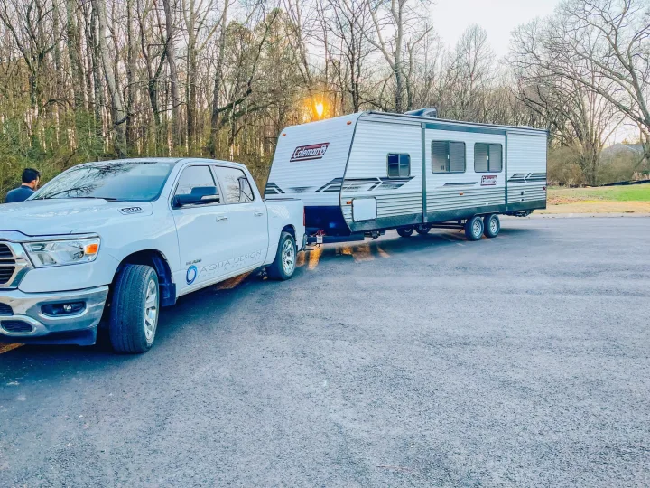 White Ram pickup truck tows a Coleman travel trailer, perfect for your rv rental adventure. Trees & sun glare in background.. Dutchmen Coleman 2022