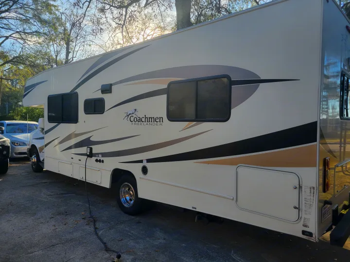 Parked Coachmen Freelander rv rental: a cream recreational vehicle with striped decals, showing a connected power cord. Ready for travel.. Coachmen Freelander 2017