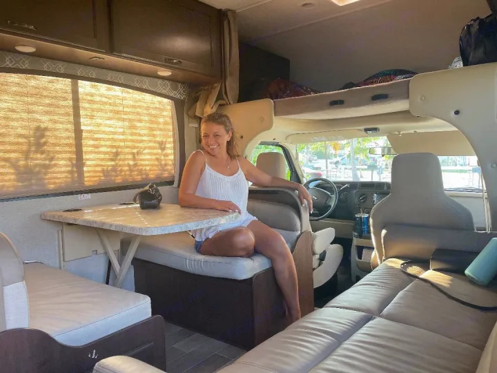 A woman sits smiling at the dinette of a brightly lit RV rental, showcasing the spacious interior and cab-over bed.. Thor Motor Coach Chateau 2020