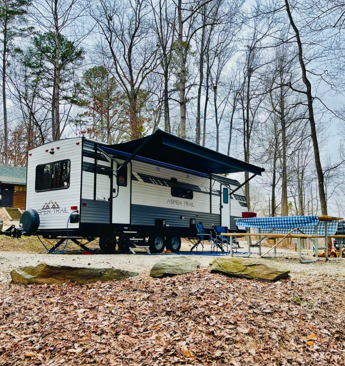 An Aspen Trail RV rental is parked at a gravel campsite with a picnic table & chairs, surrounded by bare trees & fallen leaves.. Dutchmen Aspen Trail 2022