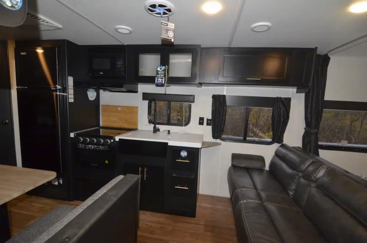 Inside this modern RV rental: a dark kitchen with a fridge, stove, and sink, plus a comfy brown leather sofa in the living area.. Forest River Cherokee 2021
