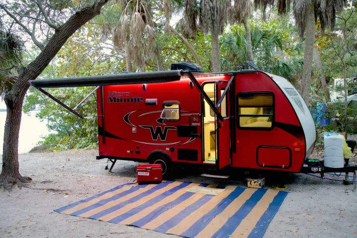Red Micro Minnie RV rental with an extended awning and striped mat, parked on sand at a lush campsite with visible water.. Winnebago Micro Minnie 2018