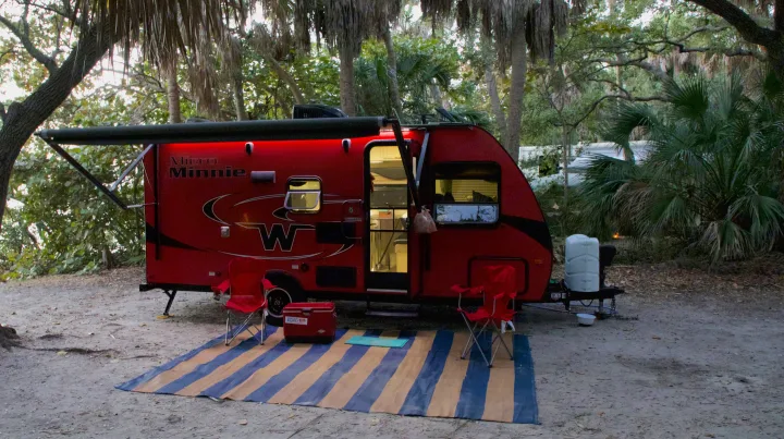 Red Micro Minnie RV rental lit up at a serene campsite with chairs and mat, perfect for a relaxing getaway.. Winnebago Micro Minnie 2018