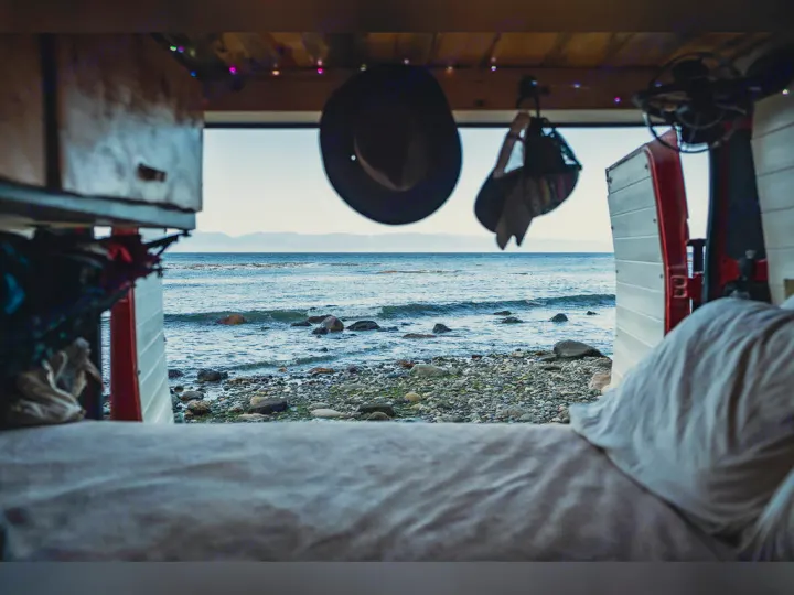 View from inside an RV rental shows a bed in foreground, beyond it a rocky beach, ocean, and distant mountains.. Dodge promaster 3500 2018