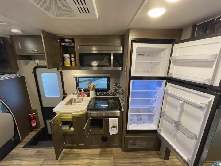 Inside this RV rental, a modern kitchen with an open refrigerator, microwave, stove, and sink is shown.. Nexus Triumph 2022