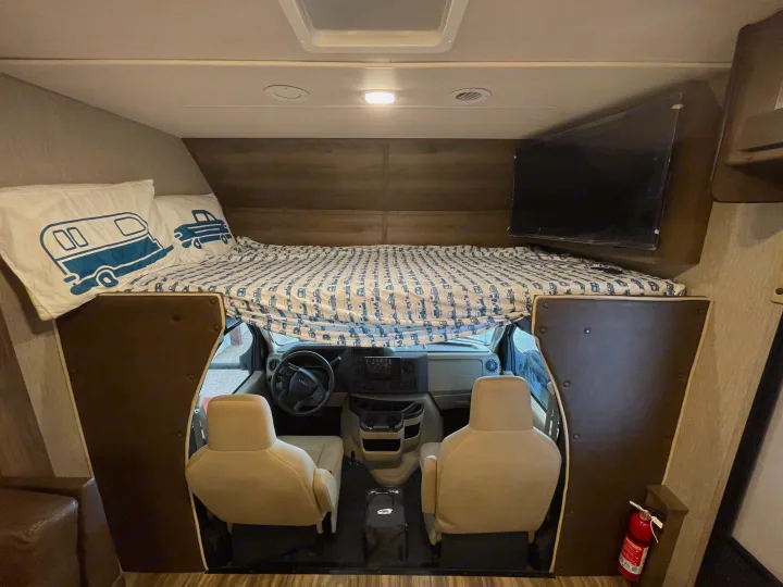 Interior of an RV rental featuring a cozy cab-over bunk bed with camper bedding, front cockpit, and mounted TV.. Nexus Triumph 2022