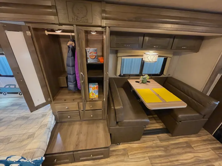 Inside this RV rental, a well-appointed interior features a wooden dinette with brown seating, a spacious wardrobe, and a bed.. Nexus Triumph 2022