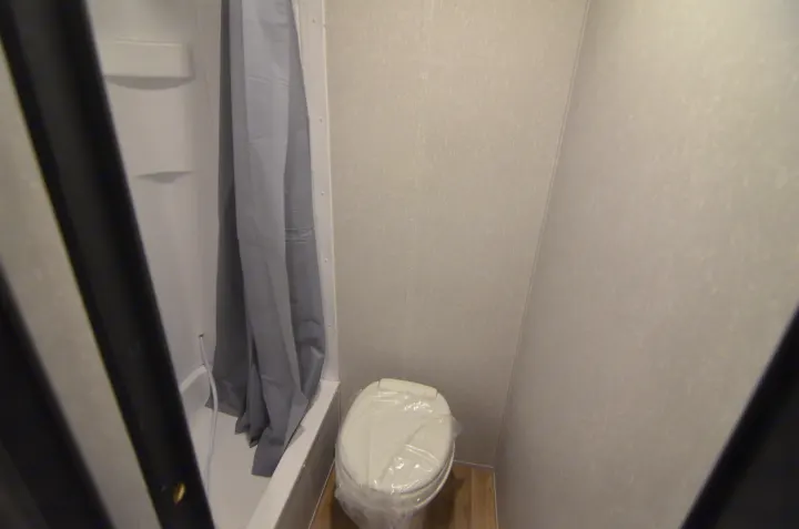 Inside an RV rental bathroom, featuring a compact shower with gray curtain and a clean, plastic-covered toilet.. Forest River Cherokee 2021