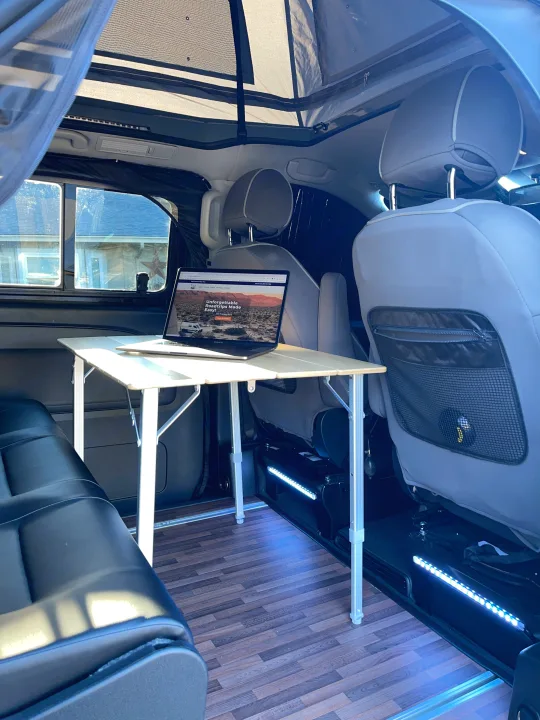Inside an rv rental: a cozy mobile workspace with a laptop on a portable table, comfy seating, and wood-look flooring.. Mercedes-Benz Metris 2020