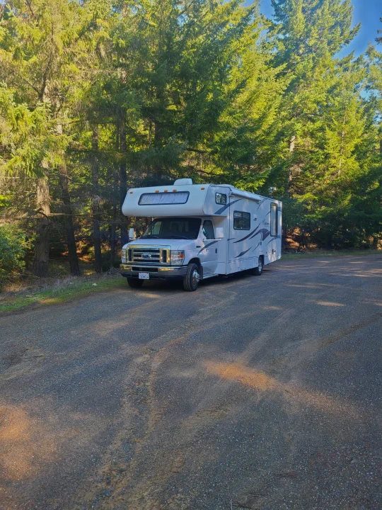 White RV rental parked on a gravel lot amidst tall green trees. Perfect for camping & travel adventures in nature.. Coachmen Freelander 2011