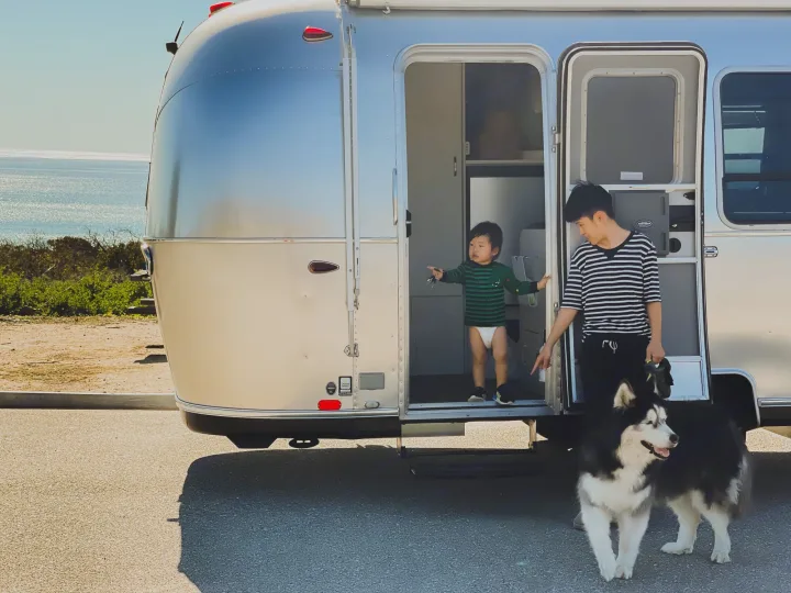 Family fun with an RV rental: an adult, a toddler peeking from their silver Airstream, and a husky-like dog beside them, ocean nearby.. Airstream Sport 2019