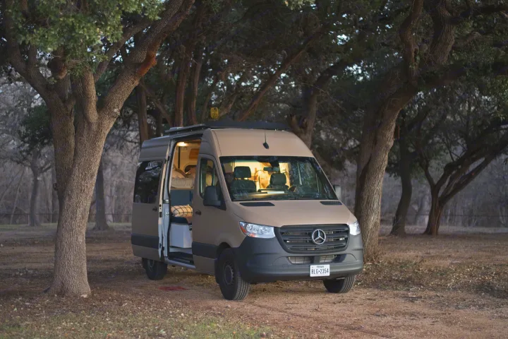 Tan Mercedes Sprinter RV rental parked amidst trees. Its open side door reveals a cozy interior, perfect for camping and outdoor adventures.. Mercedes-Benz Sprinter 2021