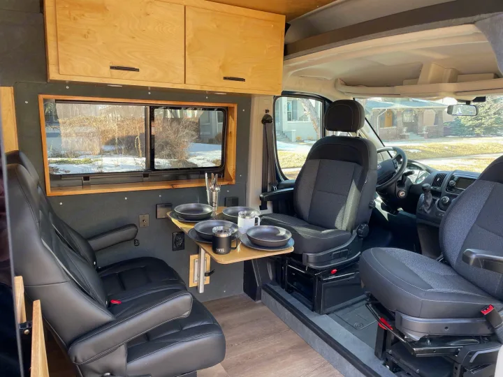 rv rental van: cozy interior with a multi-purpose dining table, swiveling seats, and wooden cabinetry by the window.. Ram Promaster 2500 2021
