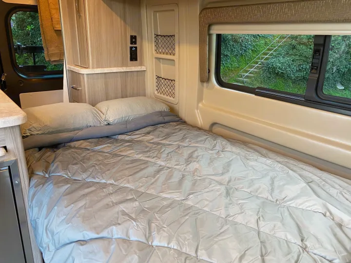 This RV rental interior features a comfortable grey bed, cabinetry, and a window showing a green, ivy-covered outdoor view.. Thor Motor Coach Scope 2022