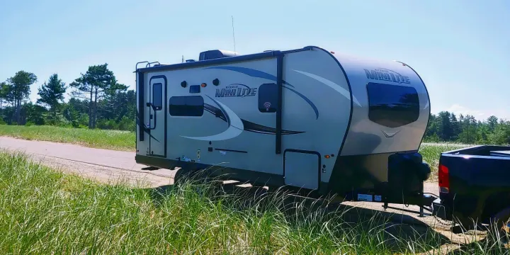 A grey travel trailer (RV rental) hooked to a black pickup truck, parked roadside with tall grass and trees under a sunny sky.. Forest River Rockwood Mini Lite 2020