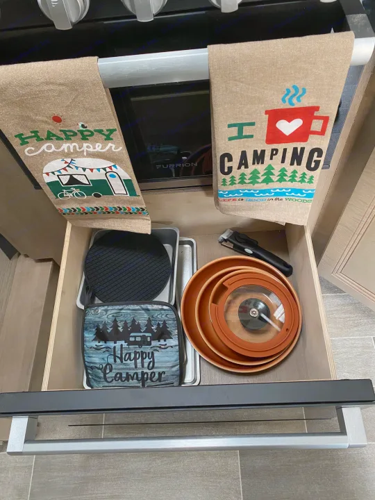 Open **rv rental** kitchen drawer with neatly arranged camping cooking essentials: pots, pans, baking trays, and "Happy Camper" items.. Thor Magnitude 2022