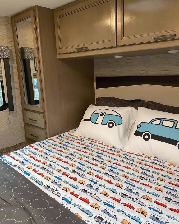 Cozy RV rental bedroom with travel-themed bedding, featuring a mirrored wardrobe, overhead cabinets, and a window.. Thor Magnitude 2022
