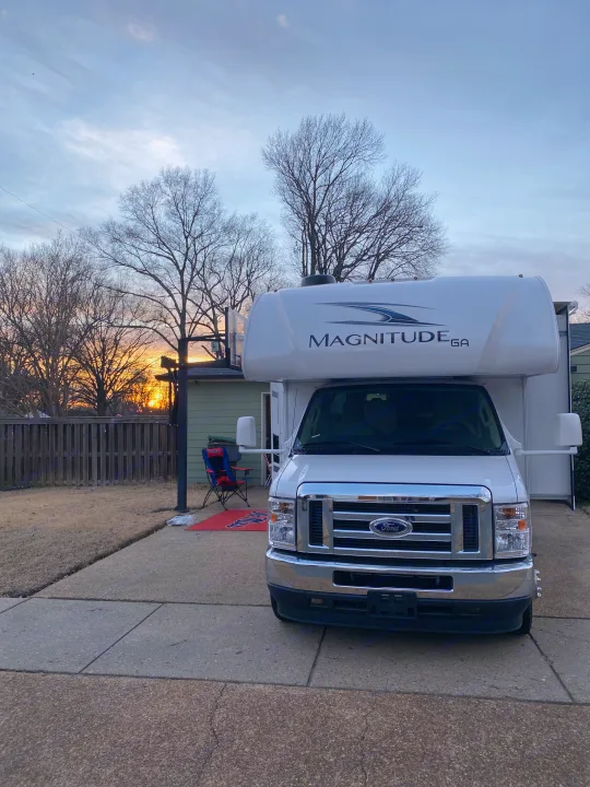 White Ford Magnitude RV rental parked on a driveway, with a beautiful sunset and bare trees in the background.. Thor Magnitude 2022