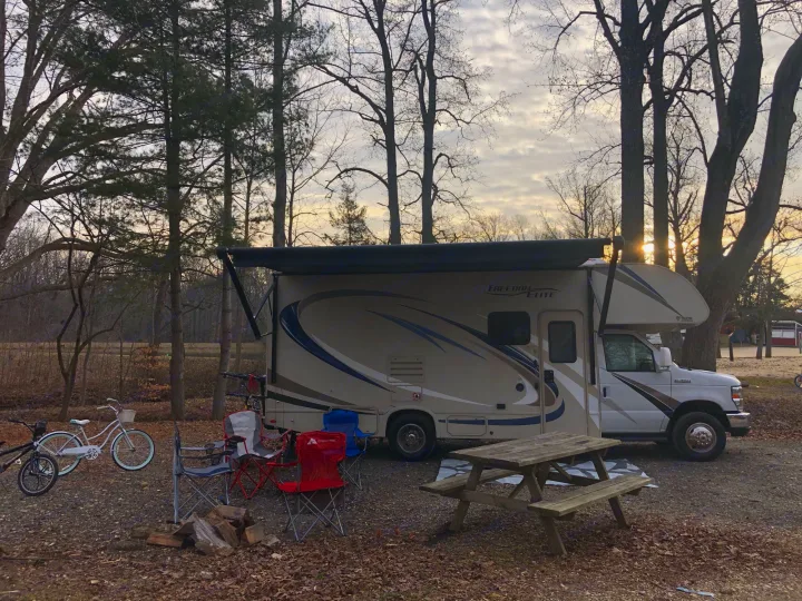 A cozy RV rental campsite at sunset. Features an RV with extended awning, chairs, bikes, and a picnic table among tall trees.. Thor Freedom Elite 2018