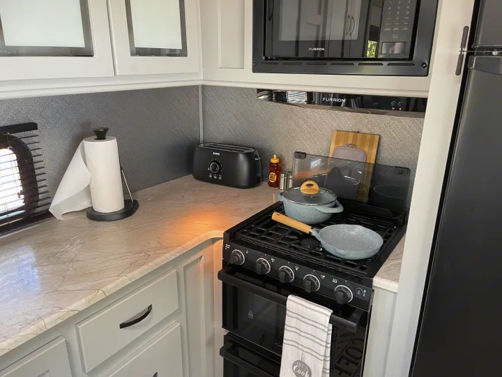 RV rental kitchen setup with white cabinets, Furrion microwave & stove, toaster, paper towels, and cookware on a marble counter.. Forest River Vibe Travel Trailer 2022