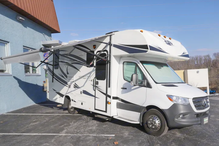 This white Jayco Melbourne RV rental motorhome is parked on an asphalt lot beside a blue building, with awnings extended.. Jayco Melbourne 2020