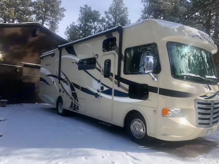 Cream-colored Class A rv rental, covered in snow & parked on a sunny winter day near a building.. Other A.C.E 2016