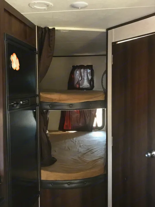 Inside an RV rental: a black refrigerator on the left and two comfortable bunk beds with dark frames and tan bedding.. Jayco Jay Flight 2018