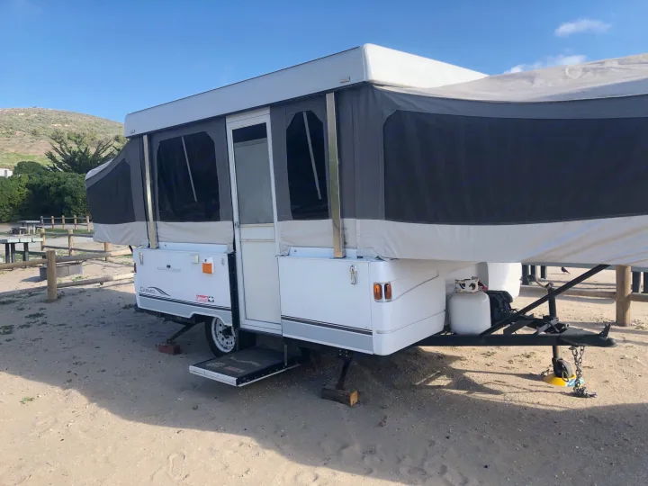 White & gray pop-up camper set up on sandy ground at a sunny campsite. Ideal for an RV rental getaway.. Coleman Grand Tour Carmel 2003