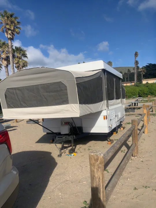 White and gray RV rental pop-up camper at a sandy campsite under a blue sky, surrounded by palm trees.. Coleman Grand Tour Carmel 2003