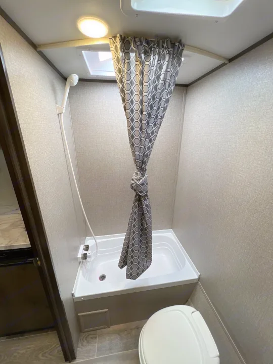 Compact RV rental bathroom with shower/tub, toilet, light, and skylight, featuring textured gray walls and a tied curtain.. Jayco Jay Flight SLX Baja Edition 2021