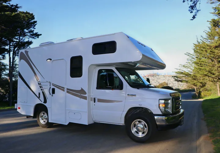 A white Class C RV rental, featuring brown graphics, is parked on a road with trees and a distant city view.. Thor Majestic 2018