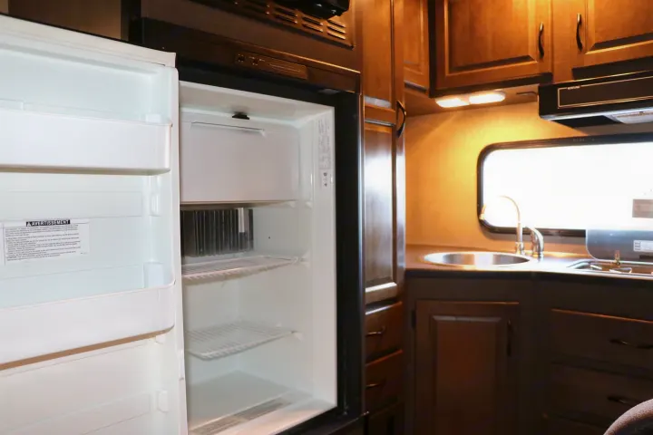 This RV rental kitchen features an open, empty refrigerator, a sink, cooktop, wood cabinets, and a window.. Thor Majestic 2018