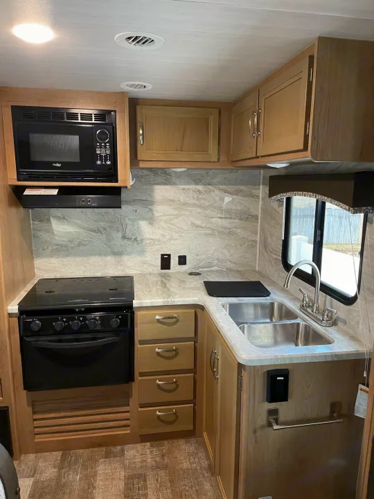 Compact rv rental kitchen featuring a modern microwave, gas stove, double sink, and light wood cabinets.. Winnebago Minnie 2020