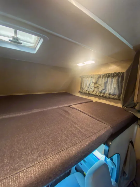 Inside an RV rental: a cozy cab-over bunk with dark mattresses, a window, overhead lights, and a roof vent.. Thor Majestic 2018
