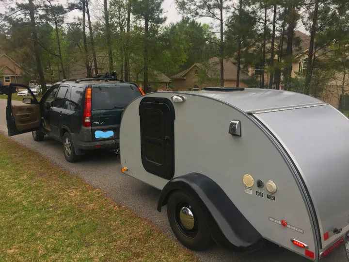 Dark green SUV with open door towing a silver teardrop camper on a paved road. Perfect for an rv rental or outdoor adventure.. Little Guy Silver Shadow 5x10 2015