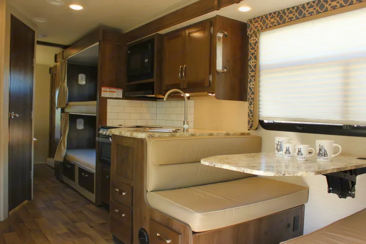 Interior of an RV rental showing kitchen area with sink, stove, and microwave, plus dinette seating, and bunk beds.. Jayco Redhawk 2020