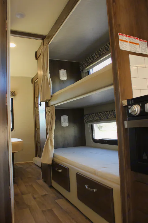 Looking into an RV rental, showing compact bunk beds with windows, storage drawers, and a partial view of the kitchen counter.. Jayco Redhawk 2020