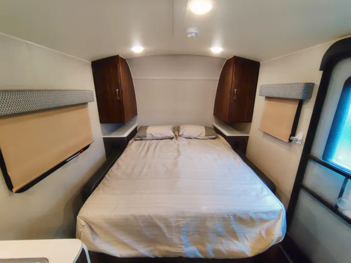 Cozy RV rental bedroom featuring a queen bed with striped bedding, overhead storage cabinets, and pull-down window shades.. Forest River Geo Pro 2018