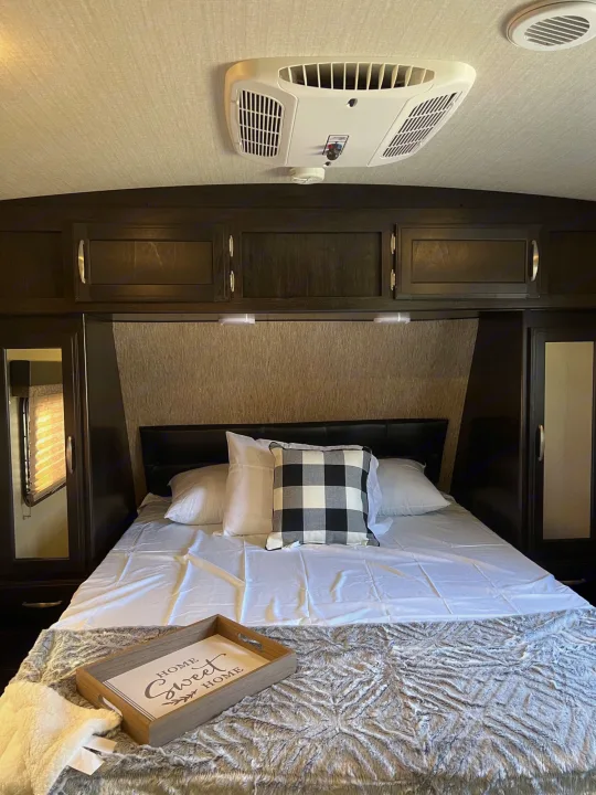 Cozy RV rental bedroom with comfortable bedding and overhead AC. Features wood cabinets and a "Home Sweet Home" tray.. Grand Design Imagine 2018