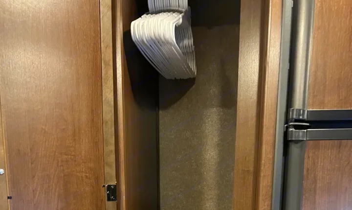 RV rental interior: an open wooden closet with a stack of white plastic hangers, beside a refrigerator.. Thor Freedom Elite 2020