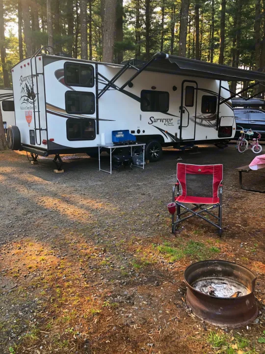 White Surveyor RV rental parked for camping with a red chair, fire pit, and grill in a gravel site among trees.. Forest River Surveyor Sport 2014