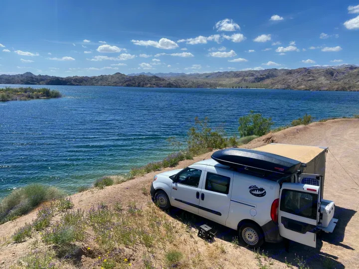 White custom camper van, perfect as an RV rental, parked on a dirt bank by a vast blue lake with mountains beyond. Ready for travel.. Cascade Campers Ram Promaster City 2020