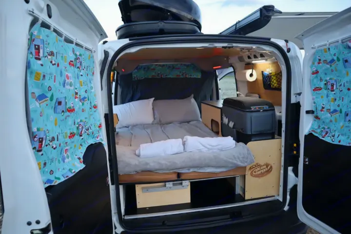 This RV rental showcases a compact campervan interior with a made bed, wood cabinets, fridge, & patterned curtains on open doors.. Cascade Campers Ram Promaster City 2020